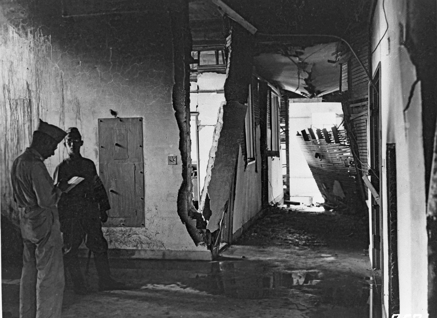 This picture shows two men standing in a house that has clearly been destroyed by the bomb, with walls askew and water damage visible. One is taking notes while the other looks on.