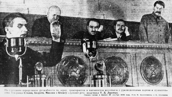Grainy photograph of Lysenko in front of microphone talking while three men are seated on podium behind him with Stalin standing and looking on.